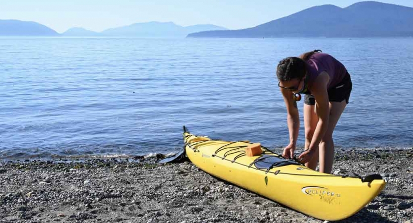 a person fixes something to a yellow kayak on the shore of a very blue lake. there are mountains in the background.
